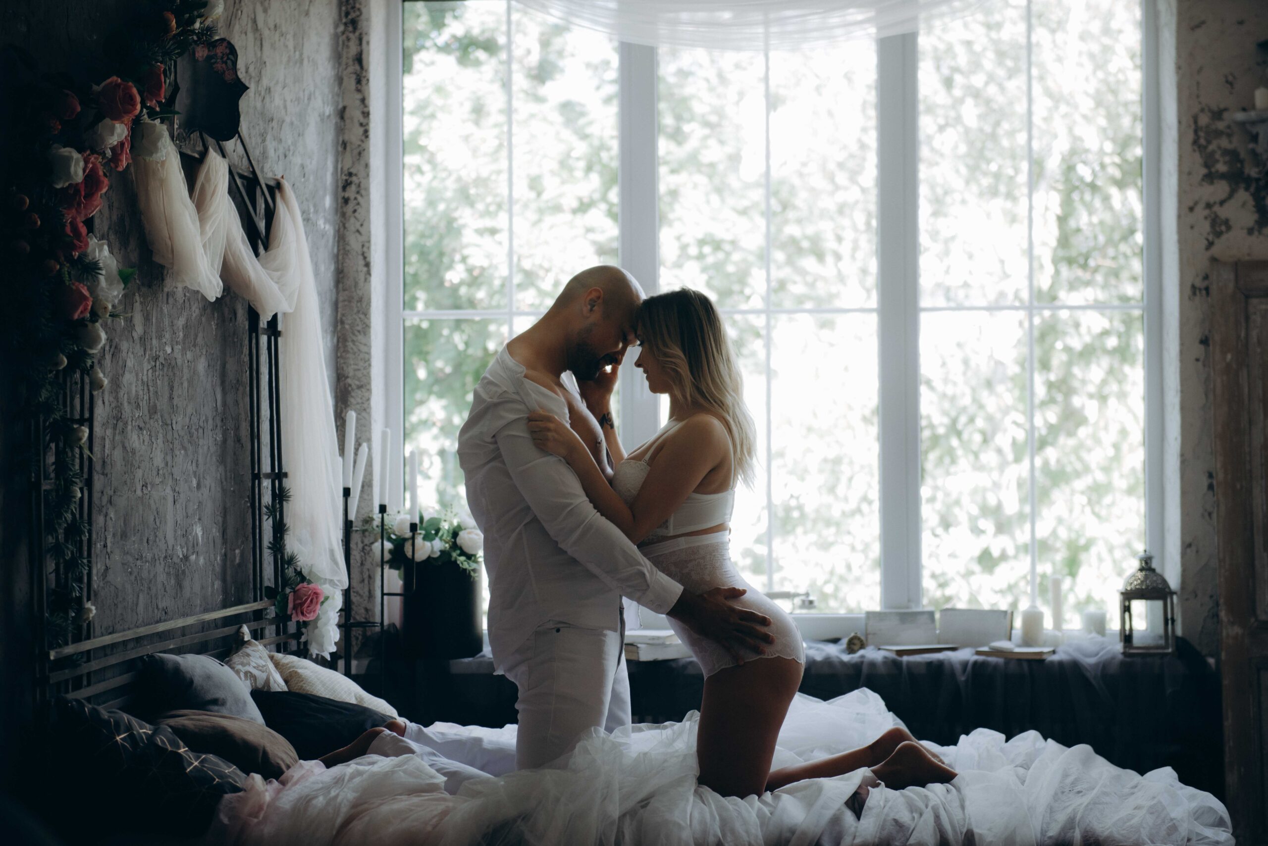 pexels-ana-maria-moroz-1700765 - Flirty Blog Ein Paar kniet auf einem Bett, im Hintergrund ist ein großes Fenster durch das man Bäume sehen kann. Sie haben die Köpfe an der Stirn zusammengelegt, sie streichelt seine Wange, er hat seine Hände an ihren Hüften.