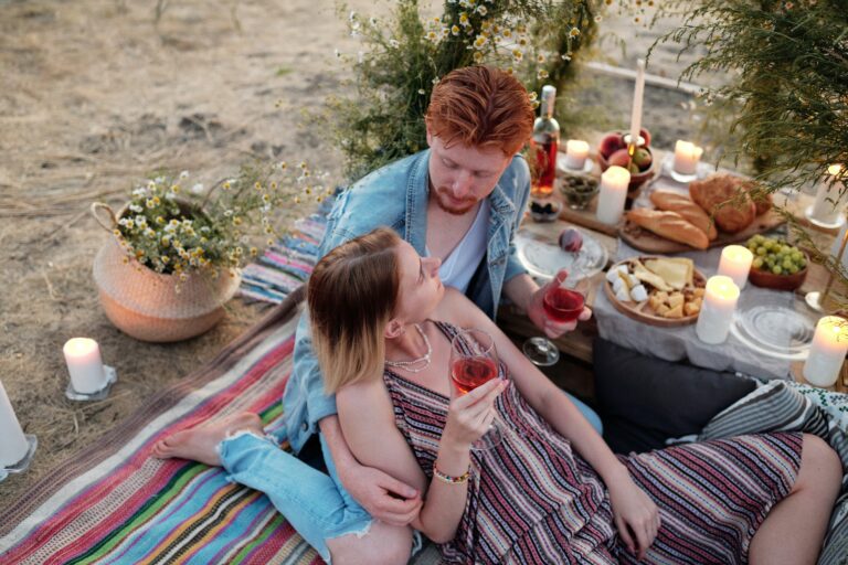 Ein junges Paar sitzt auf einer Decke am Strand, sie liegt in seinen Armen. Sie halten ein Glas Wein und auf einem Tisch sind Käse,Brot und Obst angerichtet. Einige Kerzen stehen auf dem Tisch.